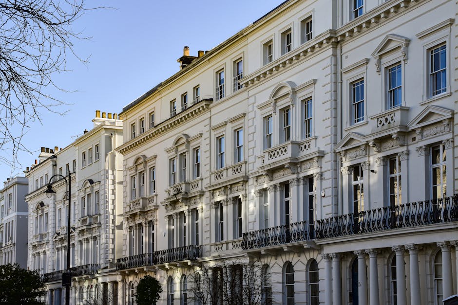 A row of white, multi-storey Georgian-style townhouses with ornate architectural details, including decorative cornices, window surrounds, and black wrought-iron balconies. The buildings feature large sash windows and are situated along a paved street with minimal street lighting, where a streetlamp is visible on the left side. The scene is captured during daylight with clear blue sky, and shadows cast by the buildings suggest the presence of early sunlight. The buildings are situated on a residential street that may be part of a home relocation or furniture transport process, with the pavement in front suitable for moving activity. External view of the properties shows currently unoccupied facades, prepared for potential interior packing or moving procedures by companies like Pimlico Movers, which specialise in removals and house moves.