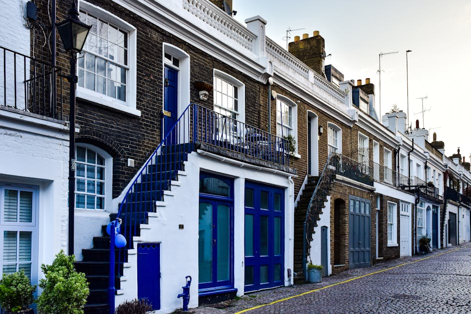 A row of terraced houses with brick facades and white window frames, featuring external staircase access to upper-floor entrances with blue-painted metal railings and stairs, situated along a cobblestone street. Each house has a ground-floor door, some with large windows, and small potted plants or bushes on the pavement. The scene appears to be during daytime with natural lighting, and the houses are closely packed, typical of a residential area prepared for home relocation or furniture transport processes, with some houses showing visible signs of packaging materials nearby. Pimlico Movers' service focus on removals is subtly supported by the depiction of a typical UK house exterior adjacent to moving and packing activities.
