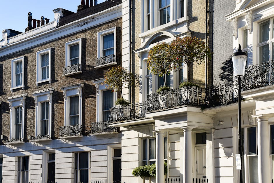 A row of white, multi-storey Georgian-style townhouses with ornate architectural details, including decorative cornices, window surrounds, and black wrought-iron balconies. The buildings feature large sash windows and are situated along a paved street with minimal street lighting, where a streetlamp is visible on the left side. The scene is captured during daylight with clear blue sky, and shadows cast by the buildings suggest the presence of early sunlight. The buildings are situated on a residential street that may be part of a home relocation or furniture transport process, with the pavement in front suitable for moving activity. External view of the properties shows currently unoccupied facades, prepared for potential interior packing or moving procedures by companies like Pimlico Movers, which specialise in removals and house moves.