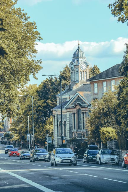 A busy street scene showing several parked cars along the curb and moving vehicles on the road, with a historic clock tower and a church building with arched windows and decorative facades in the background. To the right, part of a residential building is visible, surrounded by lush green trees and foliage. The sky is clear with scattered clouds, and the lighting suggests daytime. The scene captures elements of urban logistics and potential home relocation activities, with the environment suitable for professional removals services like those offered by Pimlico Movers to help avoid parking fines during house moves in Westminster and the surrounding area.
