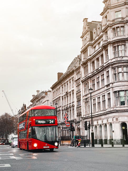 A red double-decker bus numbered 24 operated by Pimlico Movers is traveling along a street in London, passing ornate white Victorian-style buildings with large windows and decorative facades. The bus displays a route sign indicating it is heading toward Pimlico. In the background, there is a construction crane visible on the left side, and nearby, a cyclist is riding on the pavement while pedestrians wait at a crossing, some standing near traffic lights and lamp posts. The street is lined with black lampposts and street furniture, and the setting appears to be during the daytime with an overcast sky. This scene illustrates an urban environment relevant to home relocation logistics, emphasizing the importance of efficient furniture transport and loading during house removals in central London, with [COMPANY_NAME] ensuring smooth moving and packing operations to help avoid parking fines during house moves.
