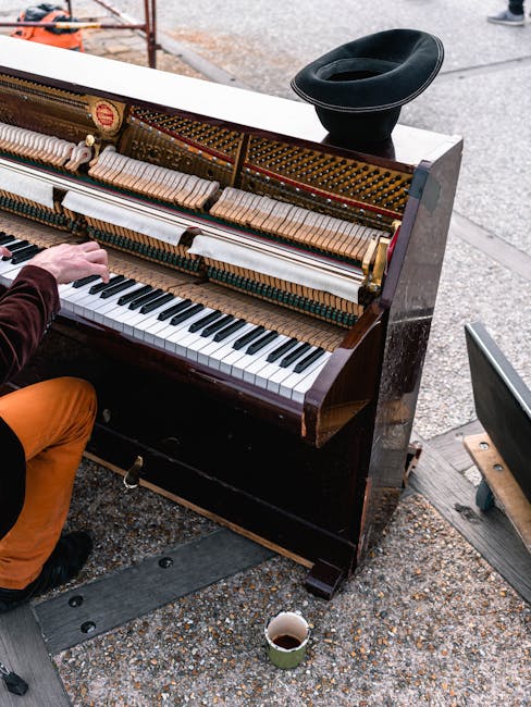 A person wearing orange trousers and a dark jacket is playing a vintage upright piano outside on a street pavement, with its top lid open to reveal the internal strings and hammers. The piano's keys are visible, showing a full range of black and white keys, and the instrument appears to be in the process of being moved or transported, as evidenced by a black trolly underneath part of the piano. An open cardboard box with packing materials rests beside a black cap placed on top of the piano, and a paper cup filled with a dark beverage is positioned on the ground nearby. In the background, a portion of a street, some paving stones, and a person's legs are visible, indicating an outdoor setting consistent with home relocation or furniture transport activities carried out by a removals service such as Pimlico Movers.