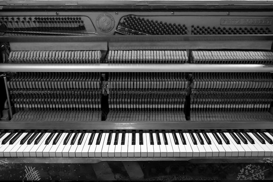 A person wearing orange trousers and a dark jacket is playing a vintage upright piano outside on a street pavement, with its top lid open to reveal the internal strings and hammers. The piano's keys are visible, showing a full range of black and white keys, and the instrument appears to be in the process of being moved or transported, as evidenced by a black trolly underneath part of the piano. An open cardboard box with packing materials rests beside a black cap placed on top of the piano, and a paper cup filled with a dark beverage is positioned on the ground nearby. In the background, a portion of a street, some paving stones, and a person's legs are visible, indicating an outdoor setting consistent with home relocation or furniture transport activities carried out by a removals service such as Pimlico Movers.