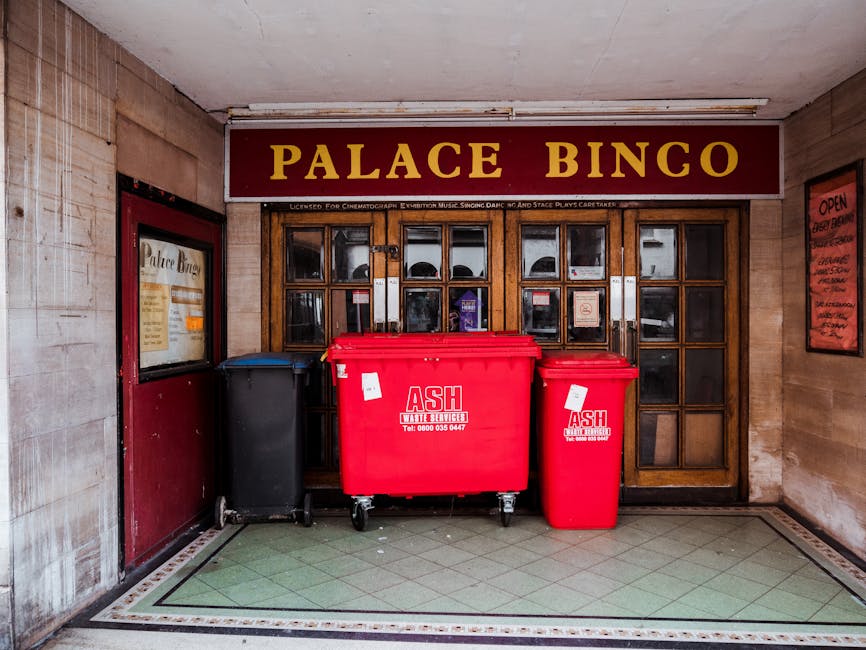 Inside the entrance area of a building with beige brick walls, featuring a wooden framed glass window and door at the back. In front of the window, there are three large wheeled bins: a black bin on the left, a large red bin in the center, and a smaller red bin on the right, all used for waste disposal. The red bins display logos with the text 'ASH Waste Services' and contact details. To the left of the black bin, there is a poster for Palace Bingo mounted on the wall, and a small window with the same signage. The floor is tiled with a pattern of green and beige squares, edged with a decorative border. The setting appears to be prepared for a home relocation or furniture transport as part of a house removal process, with items temporarily placed in the hallway before loading into a removal vehicle or van, which is not visible in the image. The overall scene reflects the logistics involved in packing, moving, and loading during a house removal by Pimlico Movers, with the environment indicating an indoor corridor or entrance lobby.