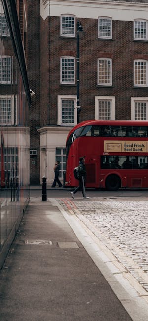 A street scene showing two individuals coordinating a home relocation outside a multi-storey brick building with white-framed windows. One person appears to be lifting or adjusting a large cardboard box wrapped in protective plastic, likely part of packing materials used during furniture transport. The second person is nearby, possibly assisting with loading or moving items. A red double-decker bus is parked by the pavement, with several windows visible and a partial advertisement on its side. The area is paved with cobblestones, and a sidewalk runs alongside the building, which has modern reflective glass windows on the left side. Visible moving equipment includes a trolley or dolly near the individuals, and the scene captures the logistics involved in executing a house removal, highlighting packing, handling, and street loading processes. Pimlico Movers, a professional removals company, may undertake such home relocation tasks efficiently in urban settings, ensuring smooth furniture transport and moving operations.