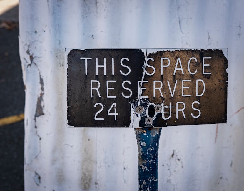 Close-up of a weathered metal parking restriction sign mounted on a blue pole, with peeling paint and rust spots. The sign reads 'THIS SPACE RESERVED 24 HOURS' in white capital letters, with some parts of the text slightly faded. It is attached to a concrete wall that shows signs of wear, dirt, and peeling paint. The image captures a typical urban setting relevant to house removals and moving logistics, highlighting the importance of complying with parking rules during home relocation or furniture transport. The sign’s worn condition reflects the potential challenges of parking enforcement in busy areas like Pimlico, where local regulations may impact moving schedules. Pimlico Movers, a professional house removals company, often coordinate with clients to navigate such restrictions to facilitate smooth packing, loading, and transportation processes.