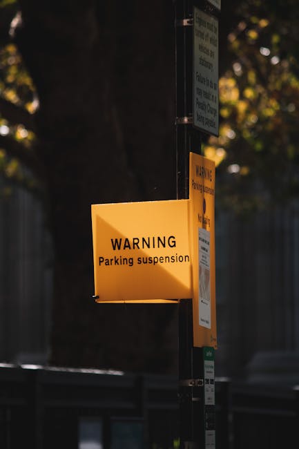 A yellow warning sign attached to a metal pole indicating a parking suspension during a home relocation process. The sign displays the text 'WARNING Parking suspension' in black, clearly visible under natural light, with a backdrop of blurred trees and a dark fence. The pole has additional smaller notices and parking information attached beneath the warning sign. This image is relevant to house removals and furniture transport services, such as those provided by Pimlico Movers, highlighting the need for permits and parking management during moving day logistics, especially in urban areas like Pimlico. The surroundings suggest an outdoor setting on a street or pavementside, with the sign positioned at a height suitable for visibility to drivers and parking enforcement personnel.