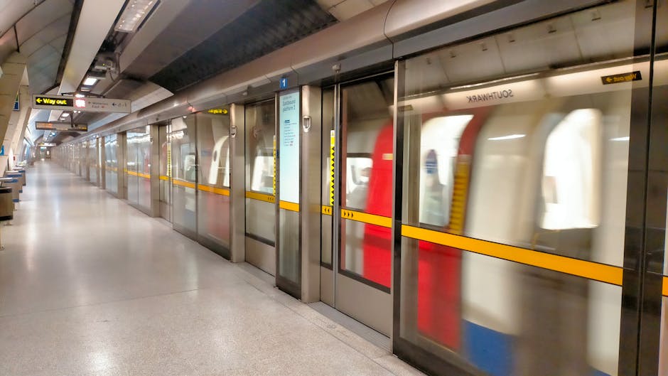 Inside the Pimlico underground station platform, the image shows a section near the staircase leading up to the street level, with a yellow 'Way out' sign hanging from the ceiling. The platform features beige tiled walls accented by brown tiles, with various informational and advertising posters mounted along the wall. A London Underground train with a red front and black windows is approaching or stationary at the platform, partially visible in the distance. The floor is made of textured tiles, with tactile paving and yellow safety lines along the edge near the tracks. To the left side of the image, metal stairs with black handrails ascend to the upper level, adjacent to a blue and white wheelchair-accessible sign. The overall lighting is dim but functional, highlighting the station's typical underground environment, which Pimlico Movers may encounter during a house relocation process involving transportation of furniture through the urban transit network.