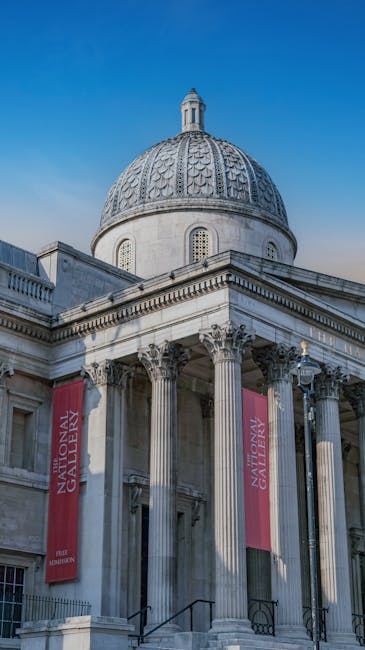 Photograph of the neoclassical National Gallery in London, featuring a grand stone facade with tall, fluted Corinthian columns supporting an entablature. The building's dome, covered in patterned tiles, rises prominently above the structure and is topped with a small decorative lantern. The exterior is illuminated by natural daylight, with a clear blue sky in the background. Red banners hanging between the columns display the name 'National Gallery.' In the foreground, a moving company such as Pimlico Movers may be involved in a home relocation process nearby, with visible cardboard boxes and packing materials possibly stored on the pavement or in the vicinity, indicating preparations for furniture transport or packing for a house move related to service pages near Tate Britain and Millbank, Pimlico.