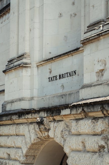 A close-up view of a historic stone building with a large, engraved sign reading 'TATE BRITAIN' on its facade, featuring detailed architectural elements such as carved stone molding and weathered surfaces. Below the sign, there is a partially visible archway with visible wear and discoloration, indicative of the building's age and exposure. The stonework shows signs of dirt and weathering, with some dark streaks and moss growth near the lower section. The photograph is taken during daylight with natural lighting illuminating the textured stone surface. Pimlico Movers, a professional removals company, may be involved in a home relocation or furniture transport process nearby, with this image capturing the building's exterior as a visual context for local moving services near Tate Britain in Pimlico, London.