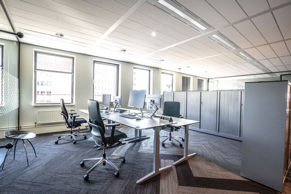 Inside a modern office space, three computer workstations with desktop monitors, keyboards, and mice are arranged on a white office table. Four ergonomic office chairs with black fabrics and adjustabl