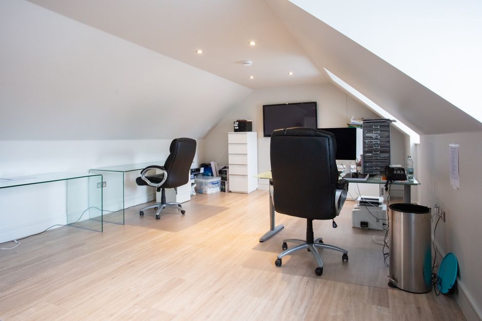 Inside a modern office space, three computer workstations with desktop monitors, keyboards, and mice are arranged on a white office table. Four ergonomic office chairs with black fabrics and adjustabl