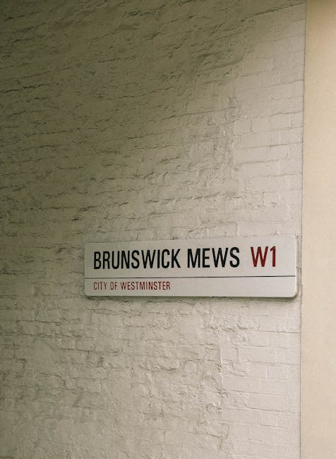 A rectangular street sign mounted on a light-colored brick wall displaying the name 'Brunswick Mews' in black text, with a red 'W1' postcode designation to the right and smaller red text underneath reading 'City of Westminster.' The sign is positioned inside a building, indicating a location in Westminster, London. The wall behind the sign has a textured brick surface, and the lighting is neutral, illuminating the area evenly, with no visible moving or packing elements in the scene. Pimlico Movers' house removals service may be involved in relocating within or through this area, supporting logistics for home or furniture transport between properties in Westminster.