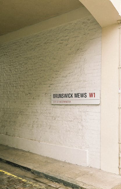A white brick wall with a street name sign reading 'BRUNSWICK MEWS W1', indicating a location within Westminster. The sign is mounted on the wall near a corner, with a small portion of a sidewalk visible at the bottom. The wall appears to be part of a building façade, illuminated by natural lighting. This setting suggests an urban environment relevant to house removals or moving services, with the street signage important for navigation and locality identification. Pimlico Movers, a professional removals company, may use this type of location during furniture transport and home relocation processes within Westminster, supporting efficient street access permissions for moving activities.